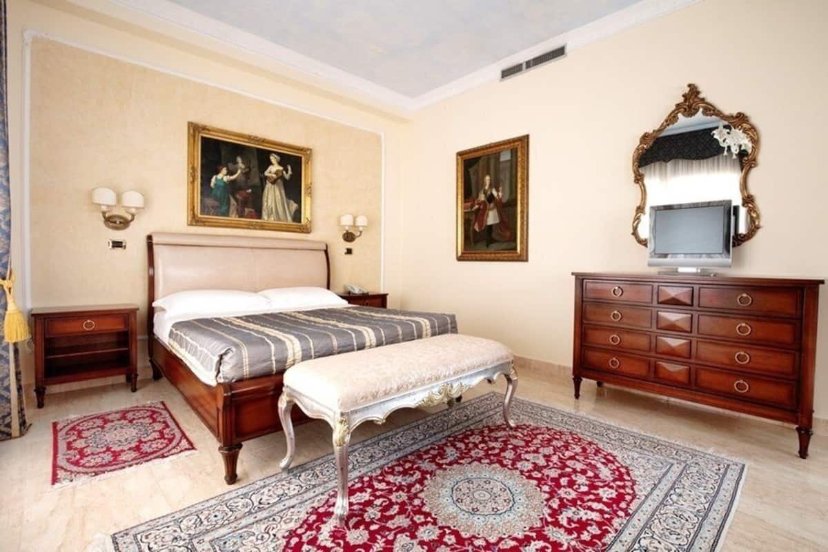 Une chambre à coucher avec un grand lit, un banc, une commode et deux lampes fixées au mur.