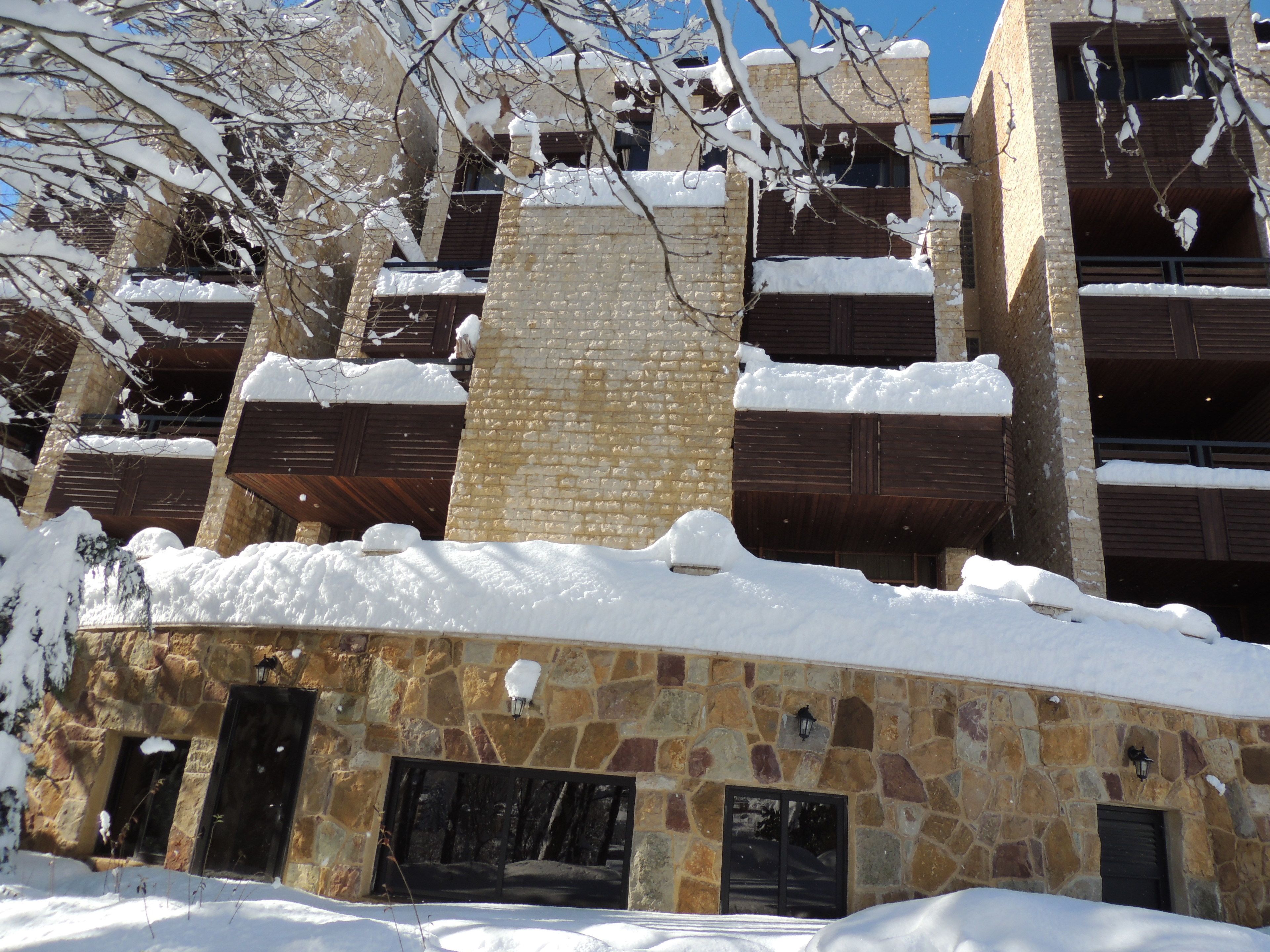 Un edificio de varios pisos con paredes exteriores de piedra y ladrillo, múltiples balcones con barandillas de madera, y acumulación de nieve en el techo y el suelo.