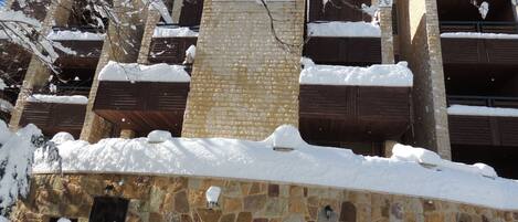 Un edificio de varios pisos con paredes exteriores de piedra y ladrillo, múltiples balcones con barandillas de madera, y acumulación de nieve en el techo y el suelo.