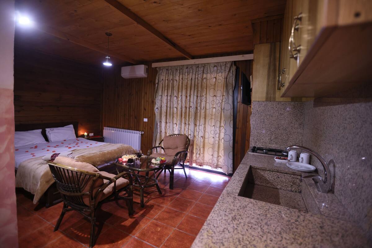 Ein gemütliches Zimmer mit Bett, Esstisch mit Stühlen, einer kleinen Küchenzeile und einem Fenster mit Vorhängen.