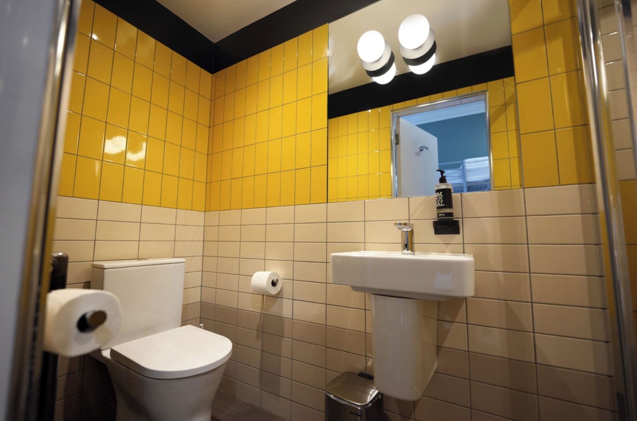 A bathroom with yellow and white tiles, a white toilet, a sink with a soap dispenser, and a mirror above the sink.