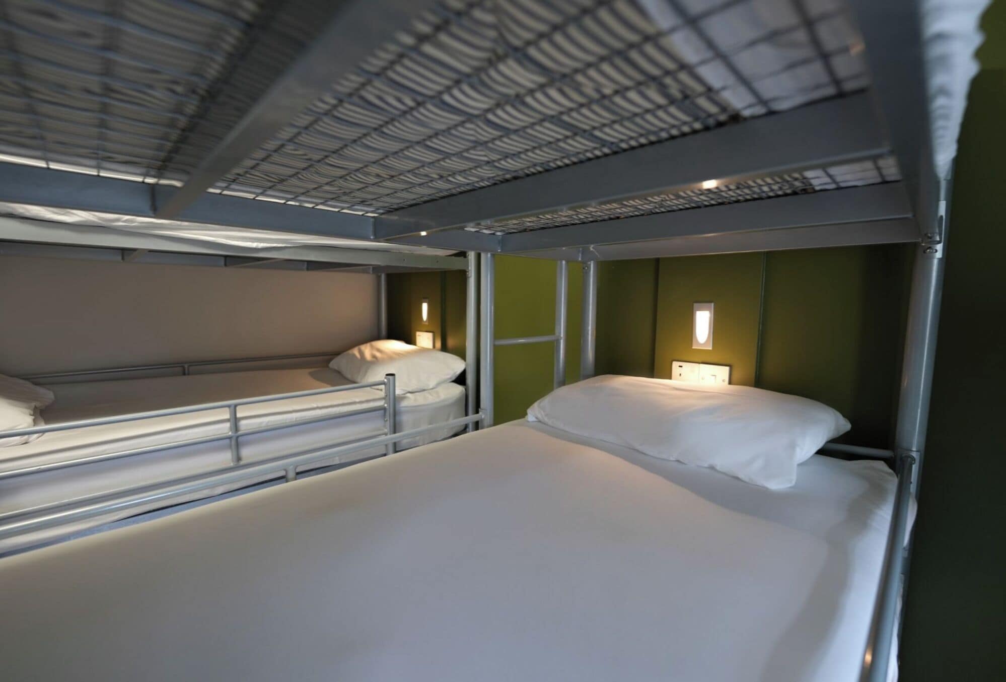 A bunk bed room with a metal frame, a green wall, and a small bedside table with a lamp.