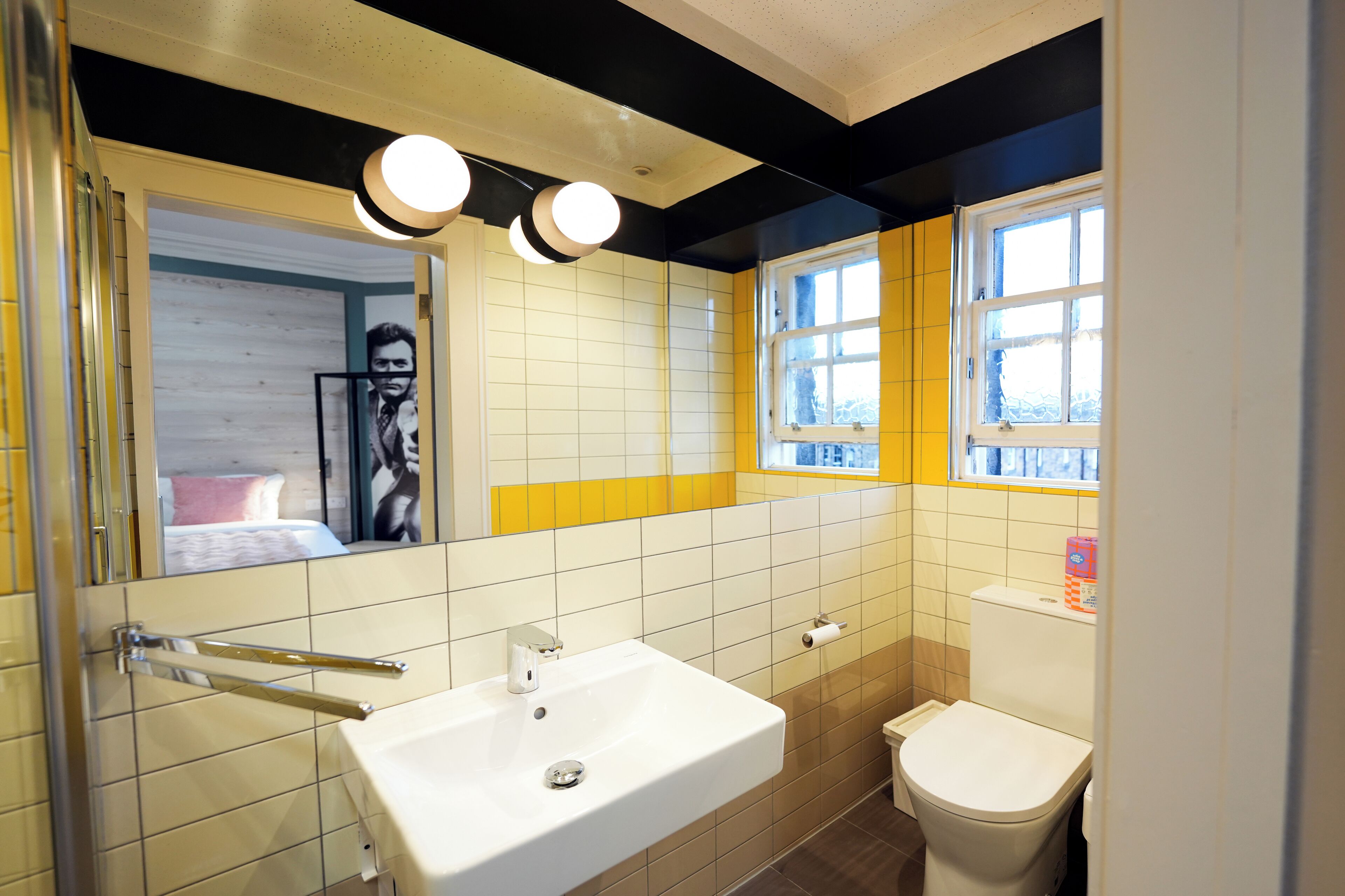 Un baño con lavamanos blanco, inodoro y espejo. Se observa una ventana con azulejos amarillos y blancos, y una puerta con un perchero para toallas.