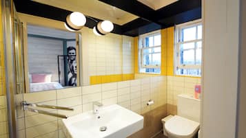 Un baño con lavamanos blanco, inodoro y espejo. Se observa una ventana con azulejos amarillos y blancos, y una puerta con un perchero para toallas.