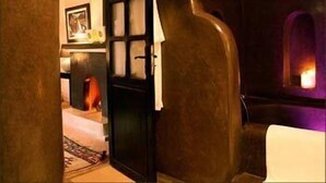 Um quarto com pouca luz, com uma grande porta de madeira escura, uma pequena janela com luz roxa e uma lareira visível.