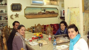 Un comedor con una mesa dispuesta para cuatro personas, rodeada de sillas. Hay un cuadro en la pared, un mapa y un reloj.