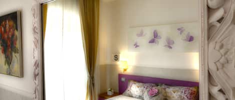 Quarto com um espelho grande, cama com roupa de cama florida, mesa de cabeceira com abajur e uma obra de arte na parede.