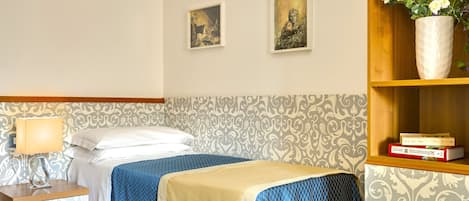 A hotel room with two beds, a nightstand, a lamp, a vase with flowers, and two framed pictures on the wall.