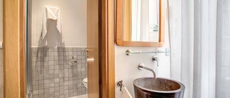 A bathroom with a glass shower enclosure, a white toilet, a wooden door, and a large, rustic sink with a woven basket.