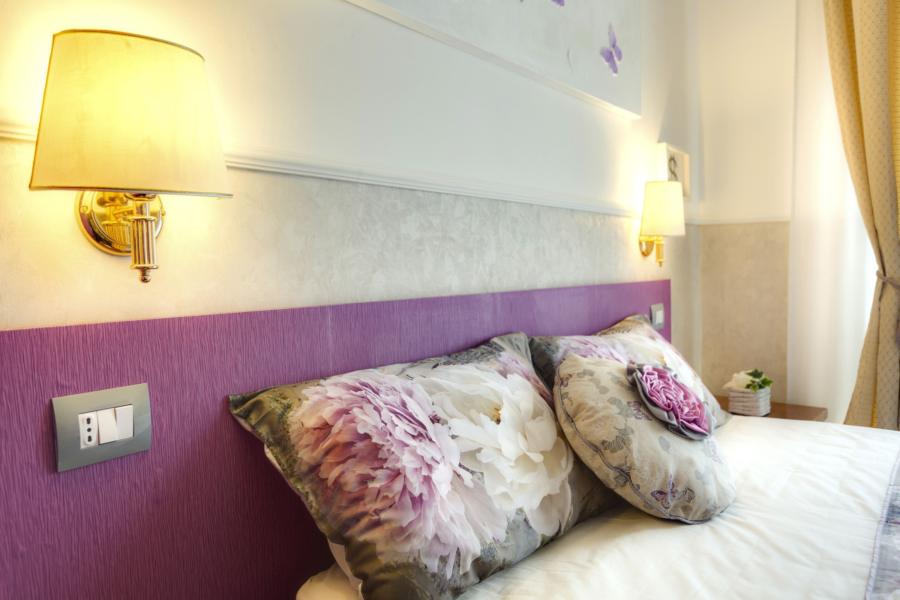Habitación de hotel con cama, dos lámparas de pared, una pared de acento morada y un cojín estampado con motivos florales.