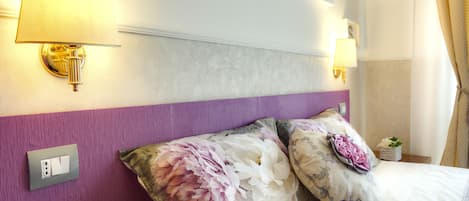 Habitación de hotel con cama, dos lámparas de pared, una pared de acento morada y un cojín estampado con motivos florales.