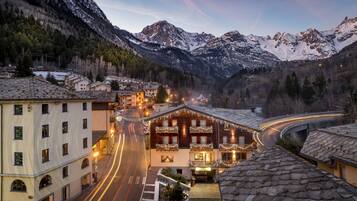 Un hotel con un edificio central que tiene balcones, rodeado de montañas nevadas y un camino sinuoso con estelas de luz.
