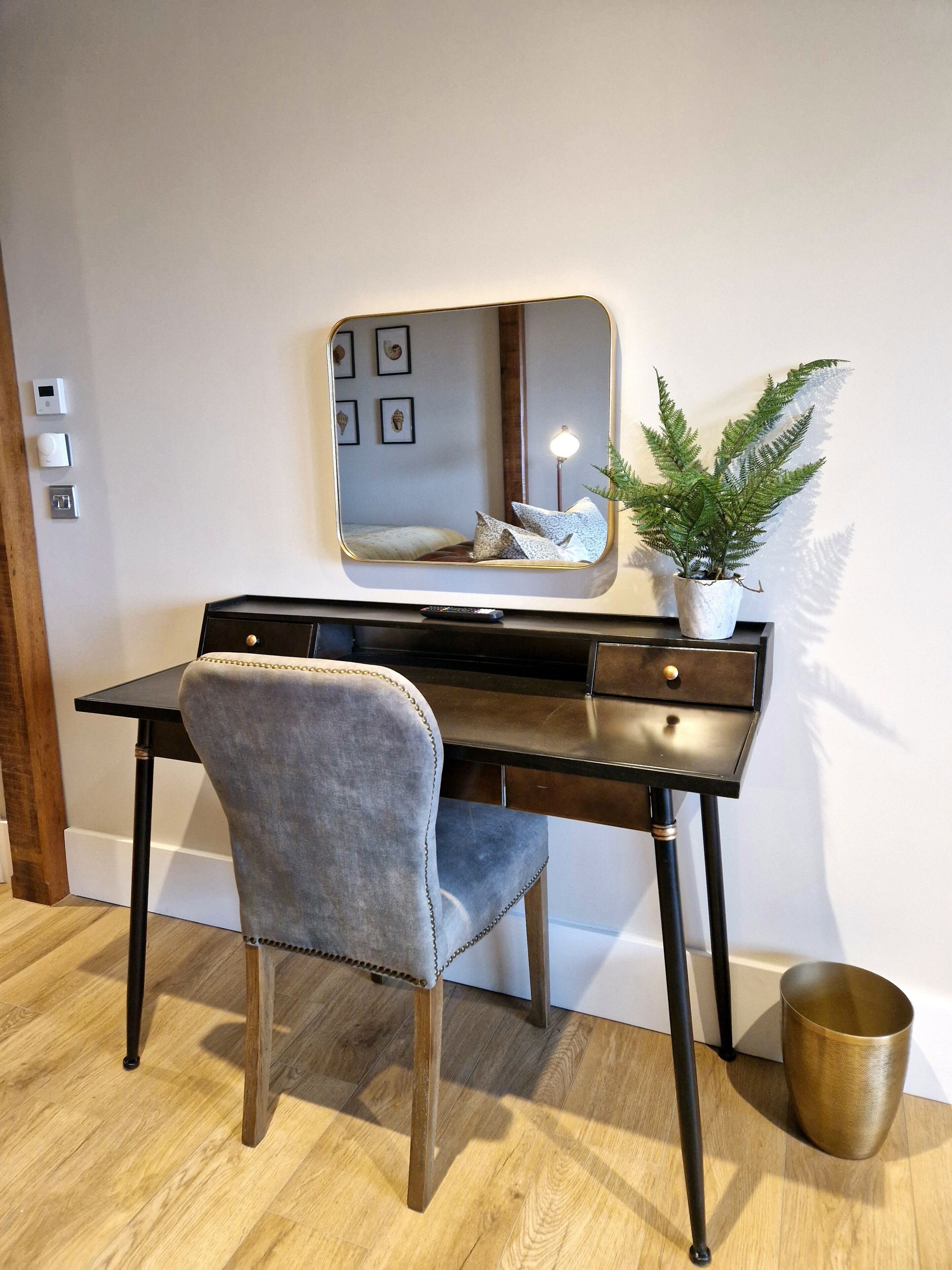 A modern office desk with a chair, a large mirror, a potted fern, and a brass trash can.