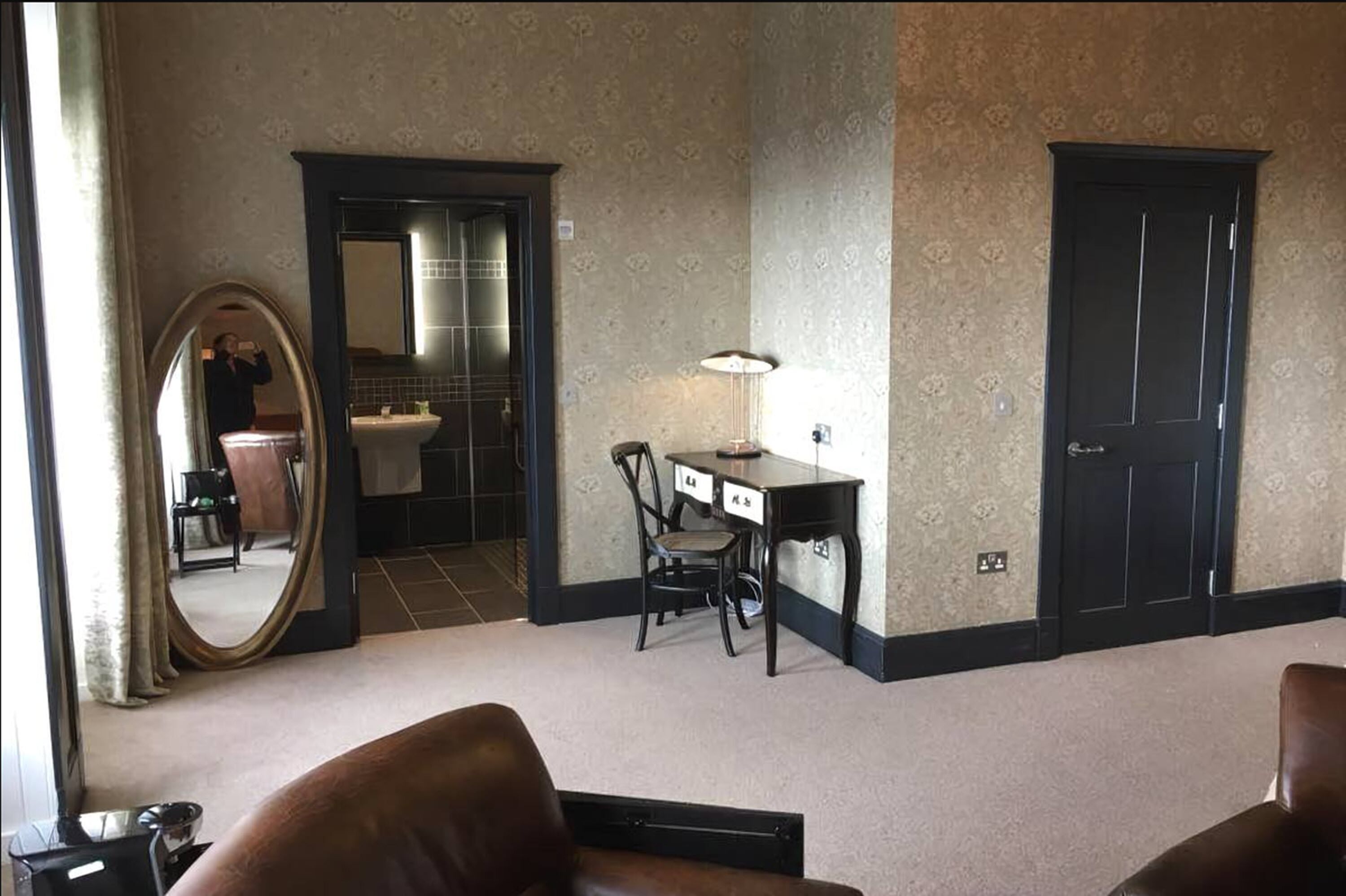 Hotellihuone, jossa on suuri peili, työpöytä tuolilla, pieni pöytä lampun kanssa ja ovi.