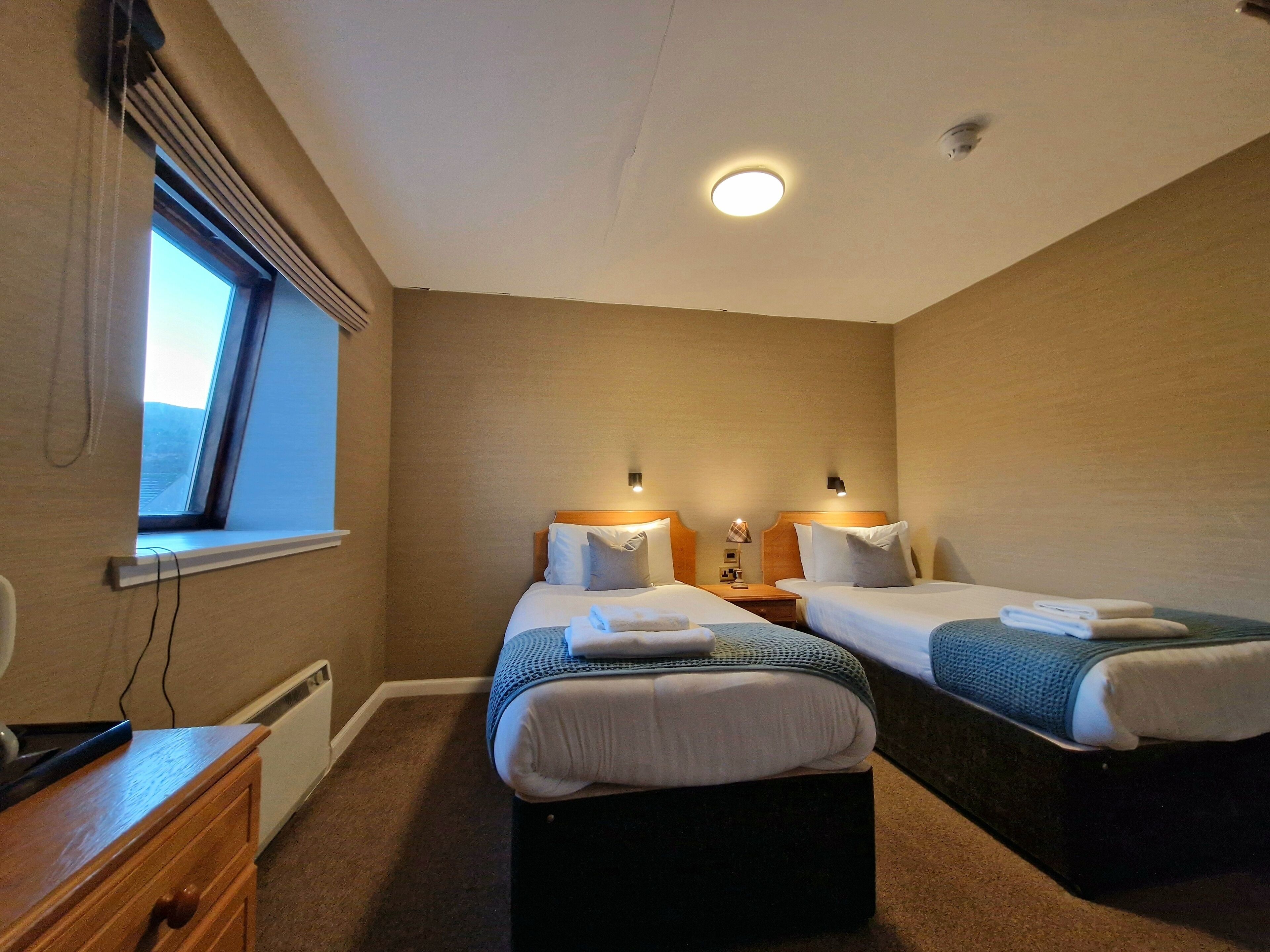 A hotel room with two beds, a window, a desk, and a radiator.
