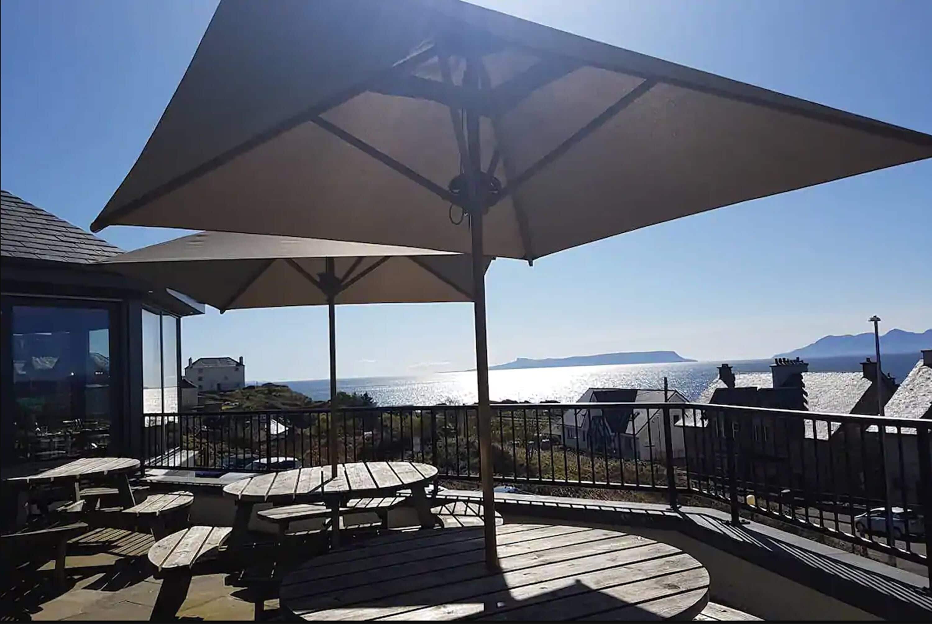 A deck with outdoor seating and a large umbrella, overlooking a scenic view of the sea and distant mountains.