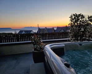 Una terraza en la azotea con un jacuzzi, una valla de madera y vistas al mar y a las montañas al atardecer.