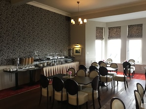 Un comedor con barra tipo buffet, que incluye recipientes con alimentos y una variedad de platos. Hay varias mesas y sillas dispuestas para los huéspedes.