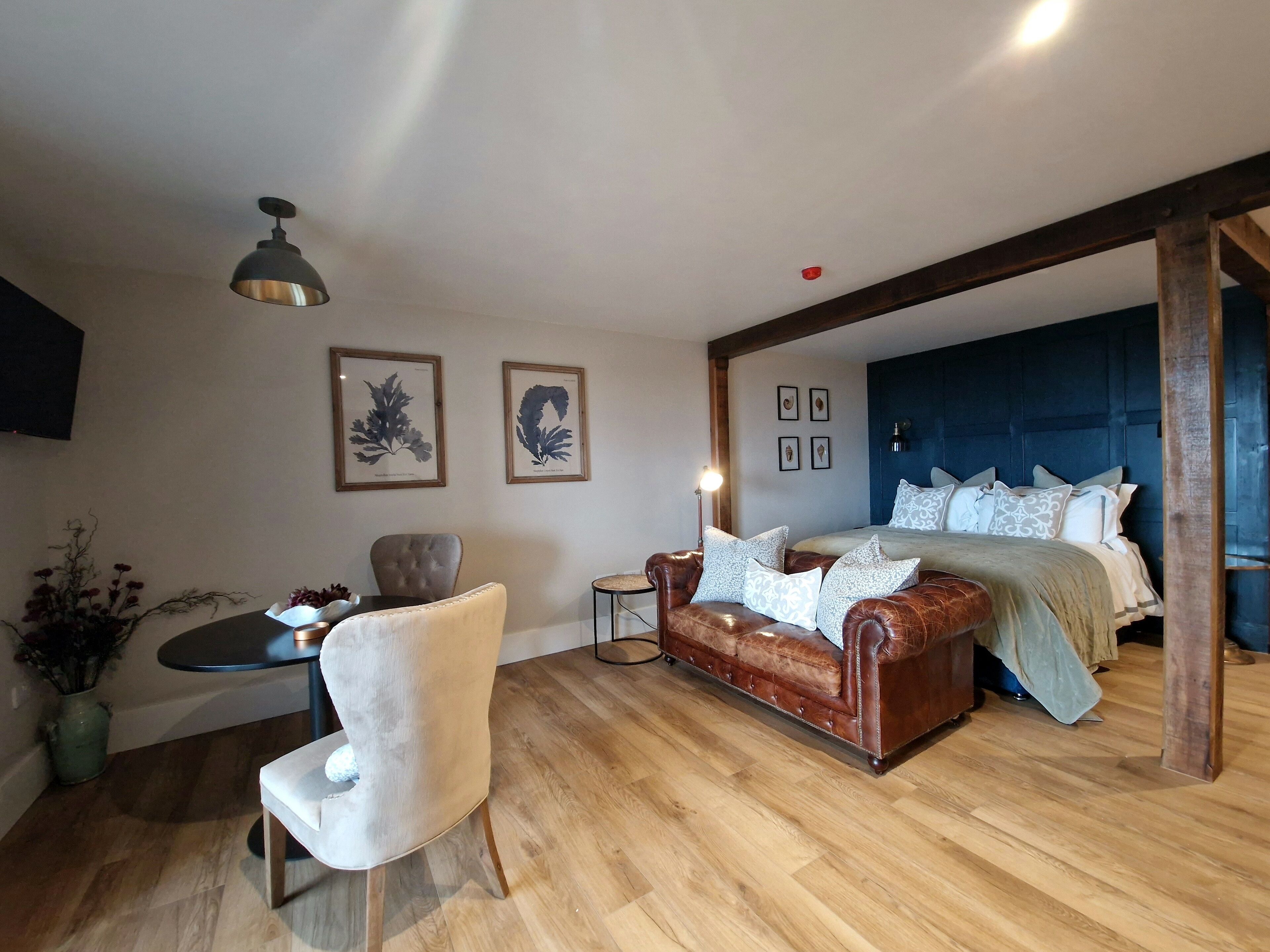 Une chambre avec un grand lit, un canapé en cuir, une petite table et deux reproductions botaniques encadrées accrochées au mur.
