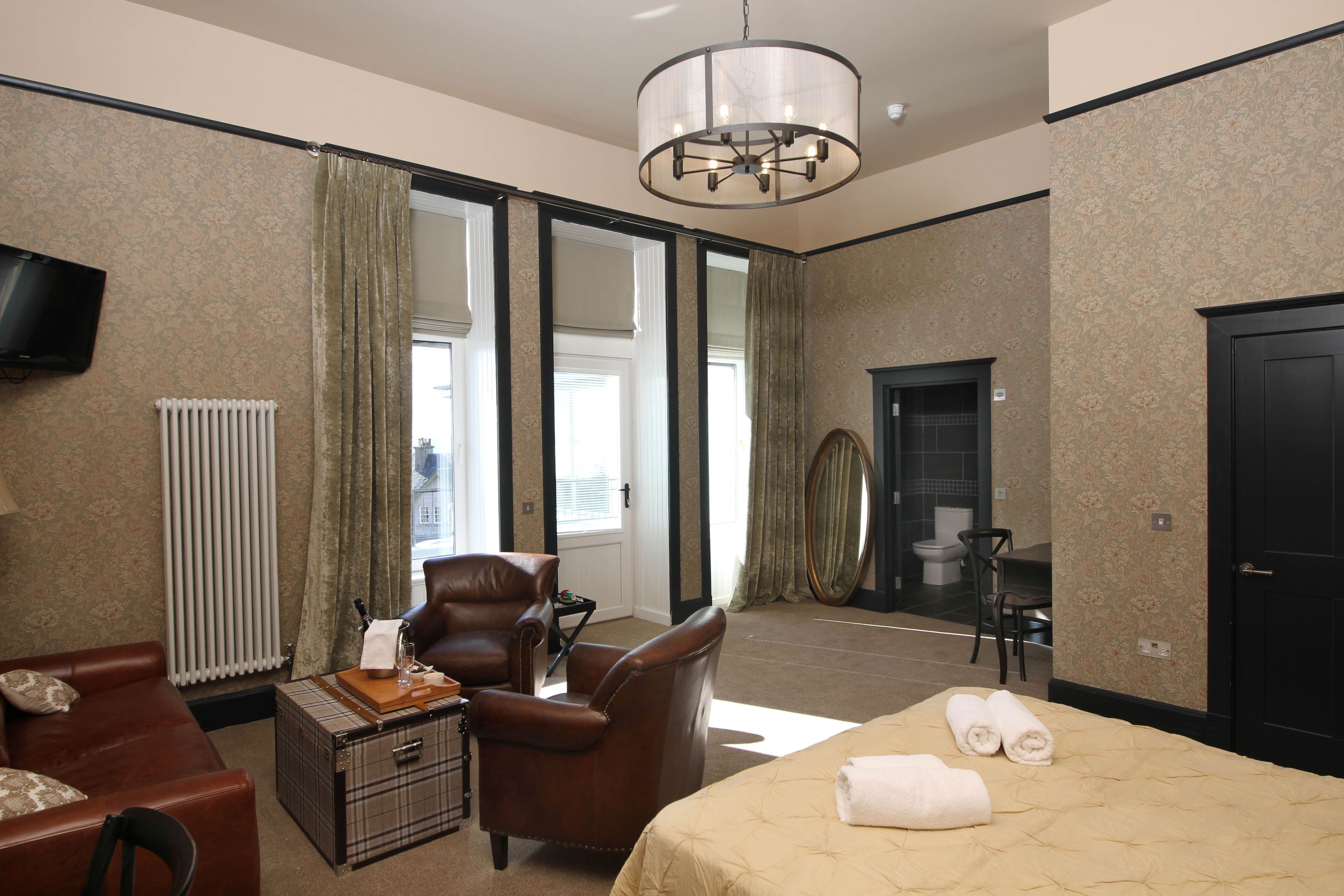 A hotel room with a bed, a leather armchair, a small table with a lamp, a television on the wall, and a view of the outside through large windows.