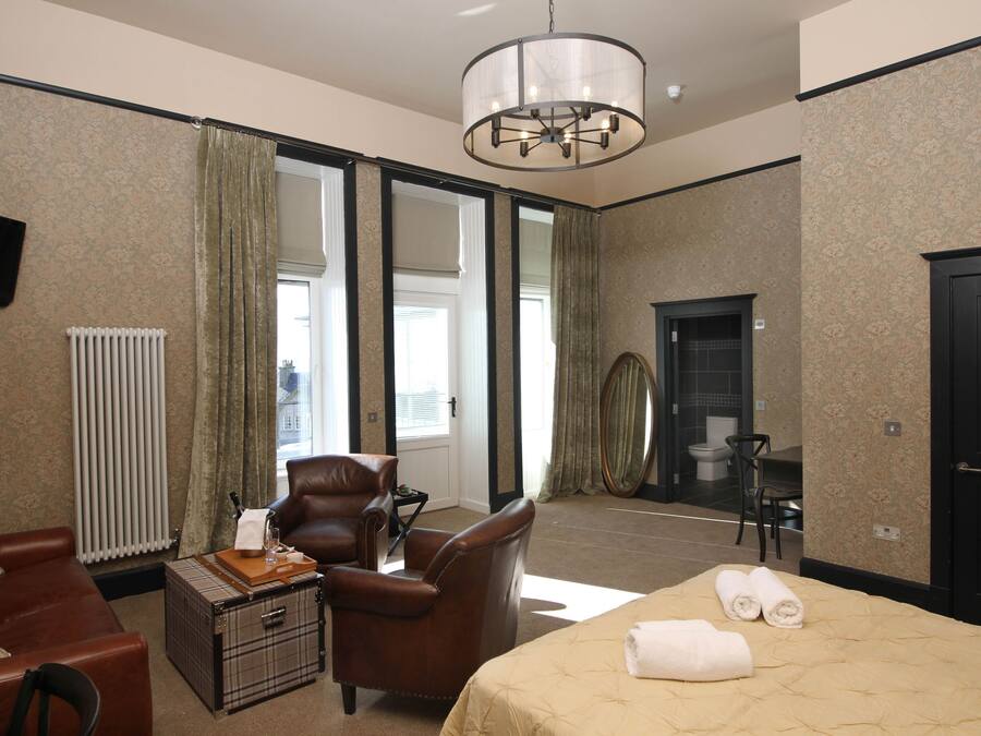A hotel room with a bed, a leather armchair, a small table with a lamp, a television on the wall, and a view of the outside through large windows.
