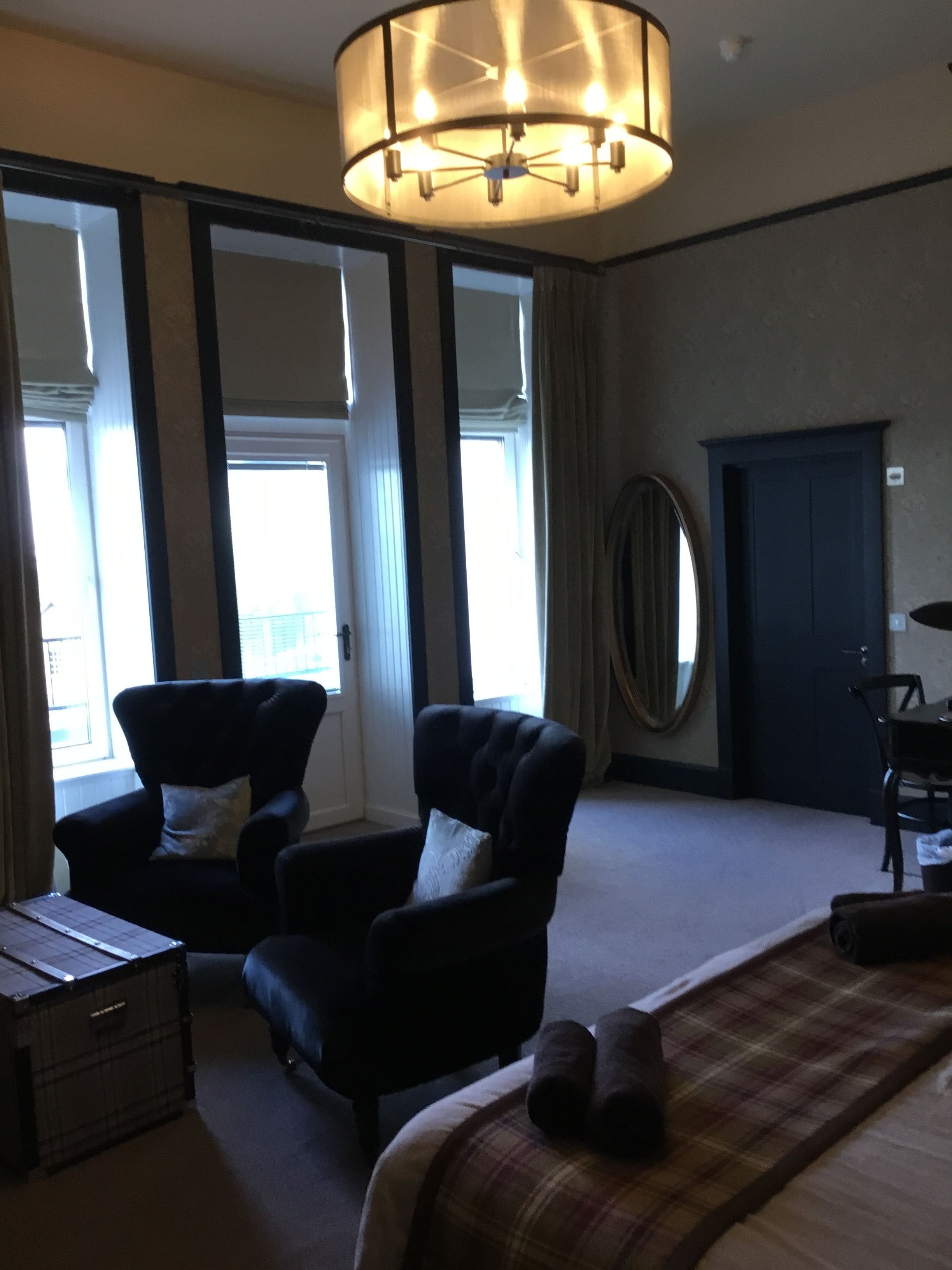 A hotel room with a large bed, two armchairs, a desk, a lamp, and a view of the outdoors through large windows.