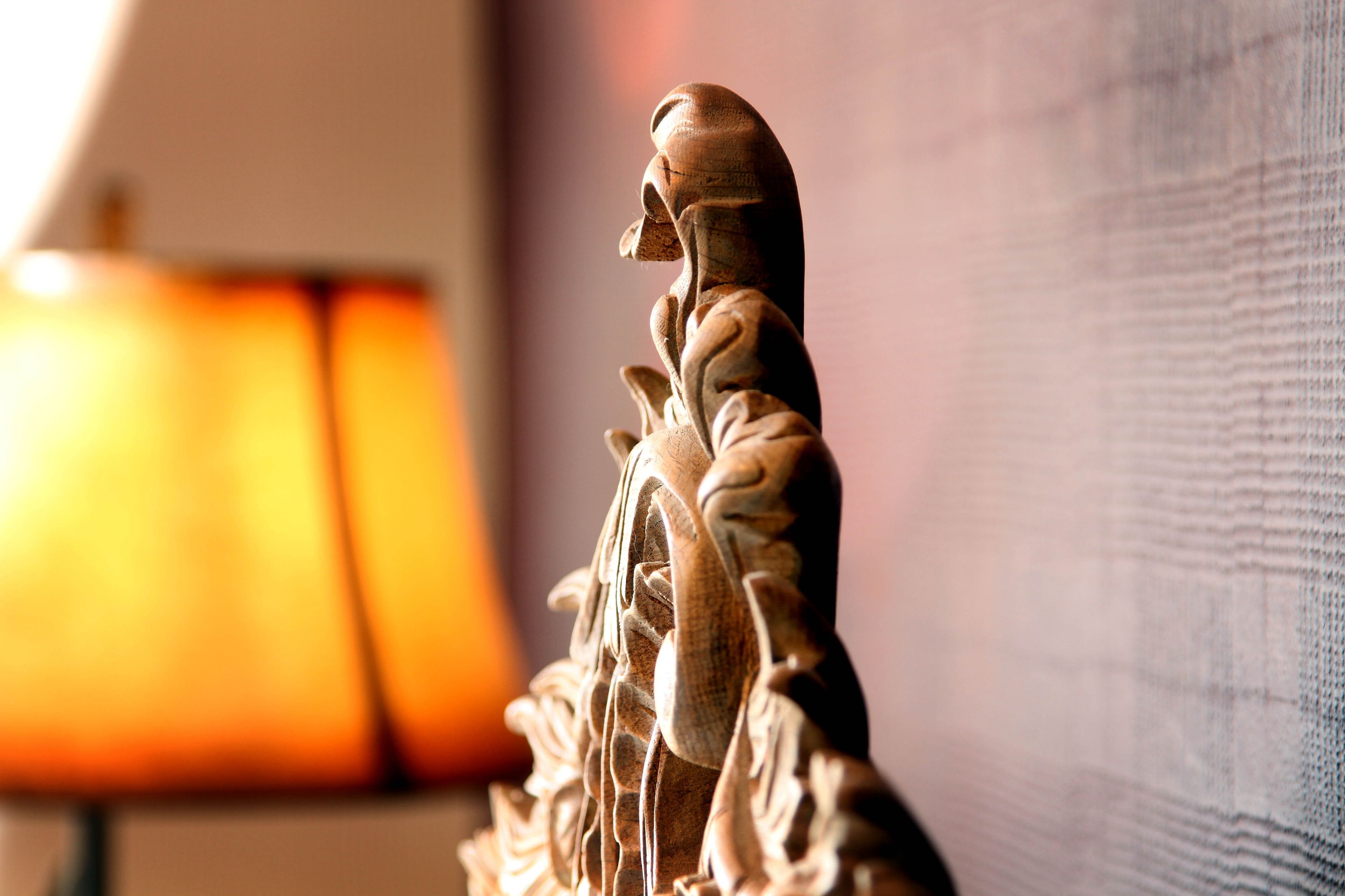 A statue with intricate carvings, positioned in a room with a blurred lamp in the background.