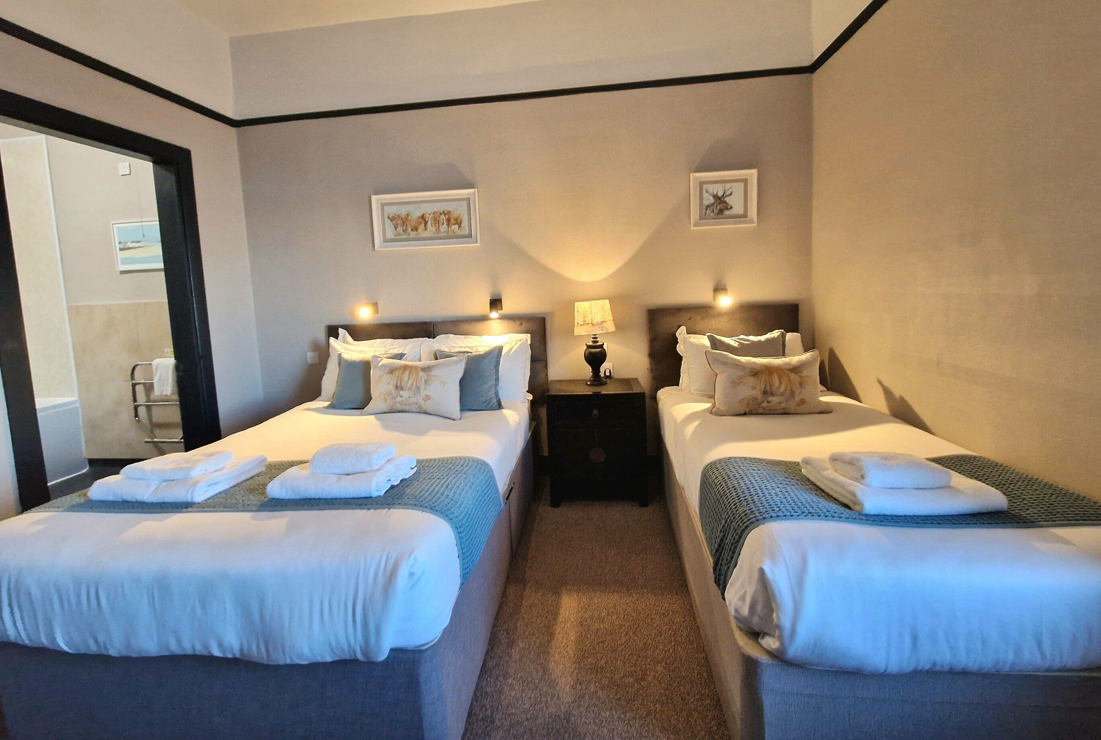 A hotel room with two beds, each with neatly folded towels and pillows. There are wall-mounted lamps and framed pictures above the beds.
