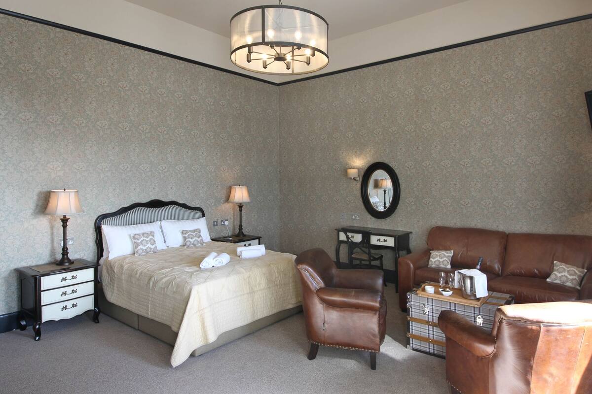 A hotel room with a bed, two bedside tables with lamps, a brown leather armchair, a brown leather sofa, a small table with a tray, and a wall-mounted TV.