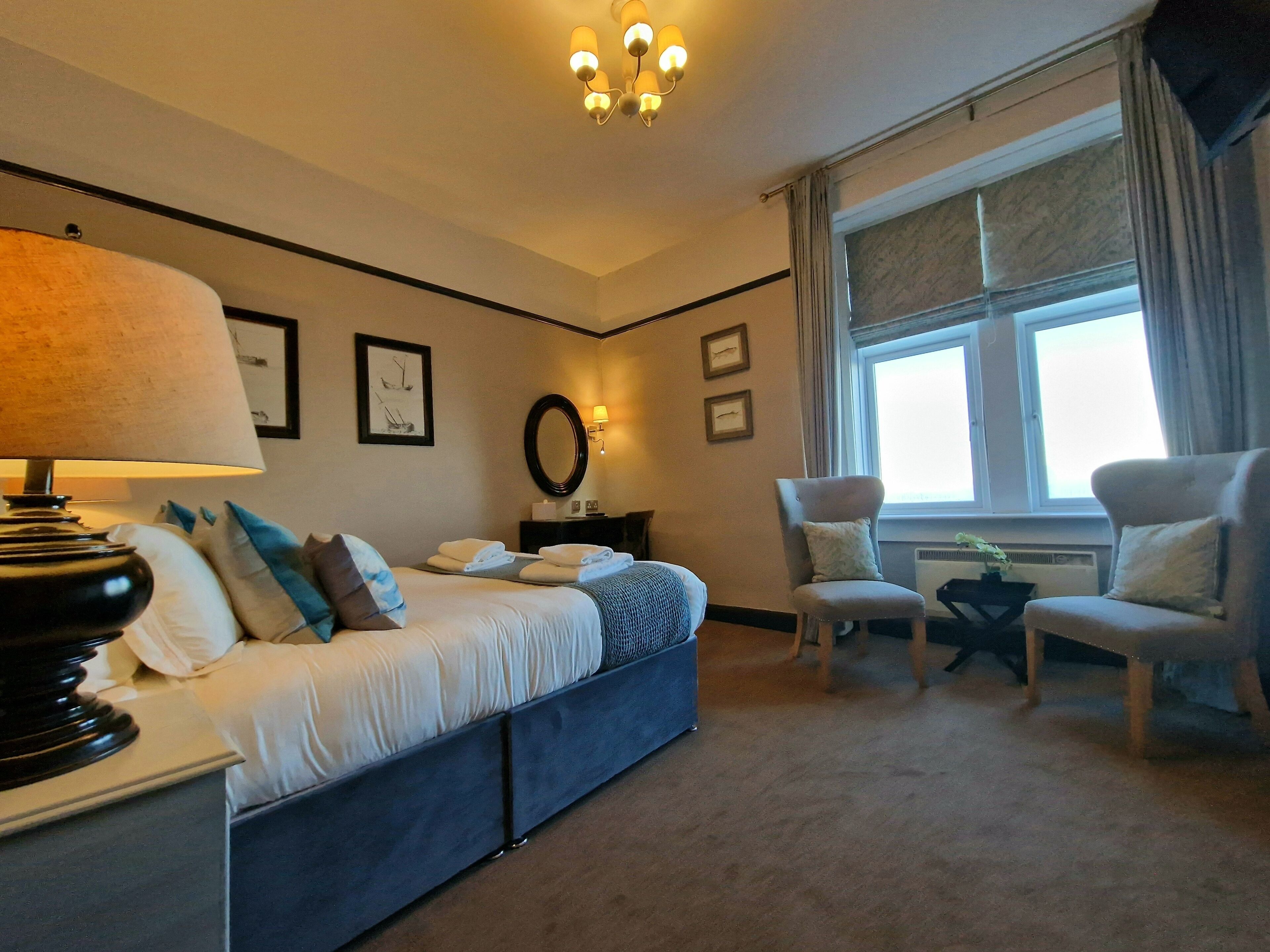 Una habitación de hotel con cama, dos sillas, escritorio, lámpara y ventana con cortinas.