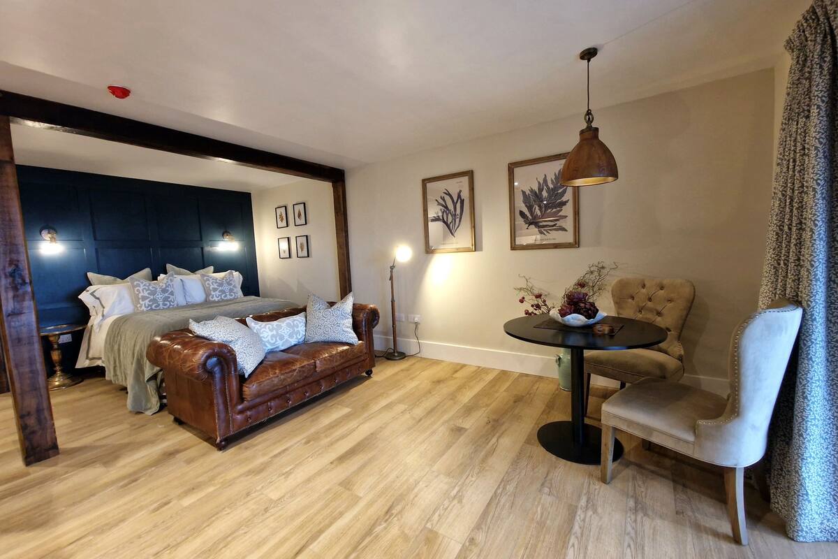 A modern living room with a leather sofa, a round table, and a chair. There is a bed with a headboard and pillows in the background.