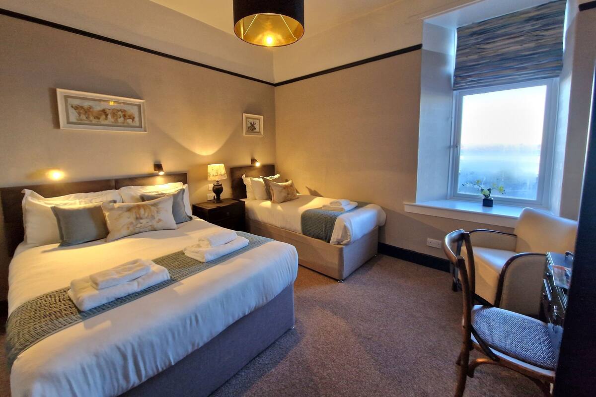 A hotel room with two beds, a desk, and a chair. There is a window with a view of the outside.