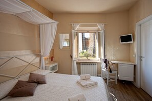 A bedroom with a bed, a desk, a chair, a TV, and a window with a view of a building. - Il Barlanzone (Montalcino)