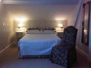 A small, dimly lit hotel room with a single bed, two bedside lamps, a chair, and a mirror on the wall.