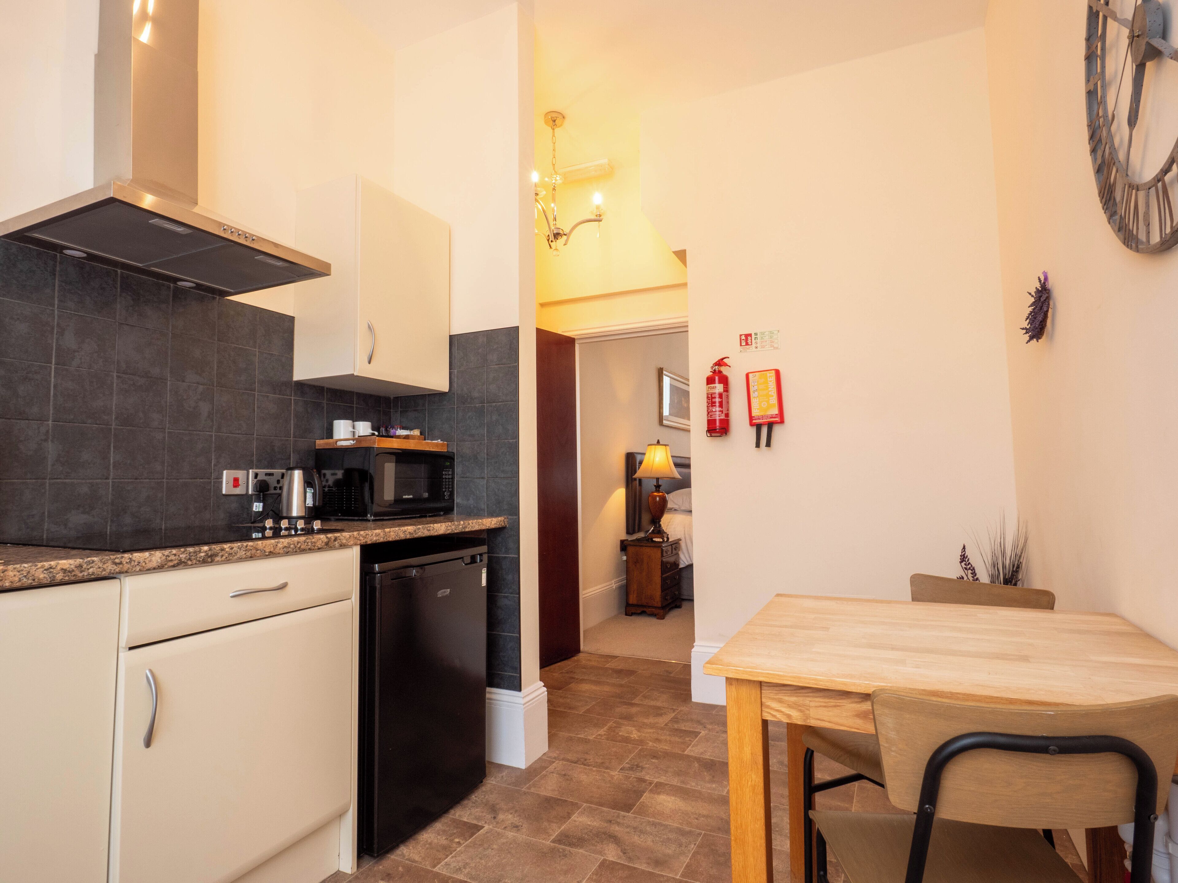 A compact kitchen with a microwave, coffee maker, and dishwasher. There is a dining area with a table and chair, and a clock on the wall.