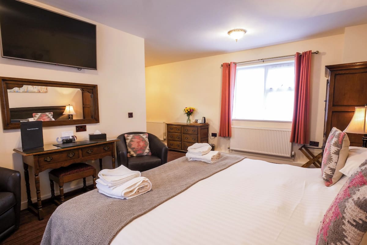 Ein Hotelzimmer mit einem großen Bett, einem Schreibtisch mit Computer, einem Sessel, einem Schminktisch und einem Fenster mit Vorhängen.