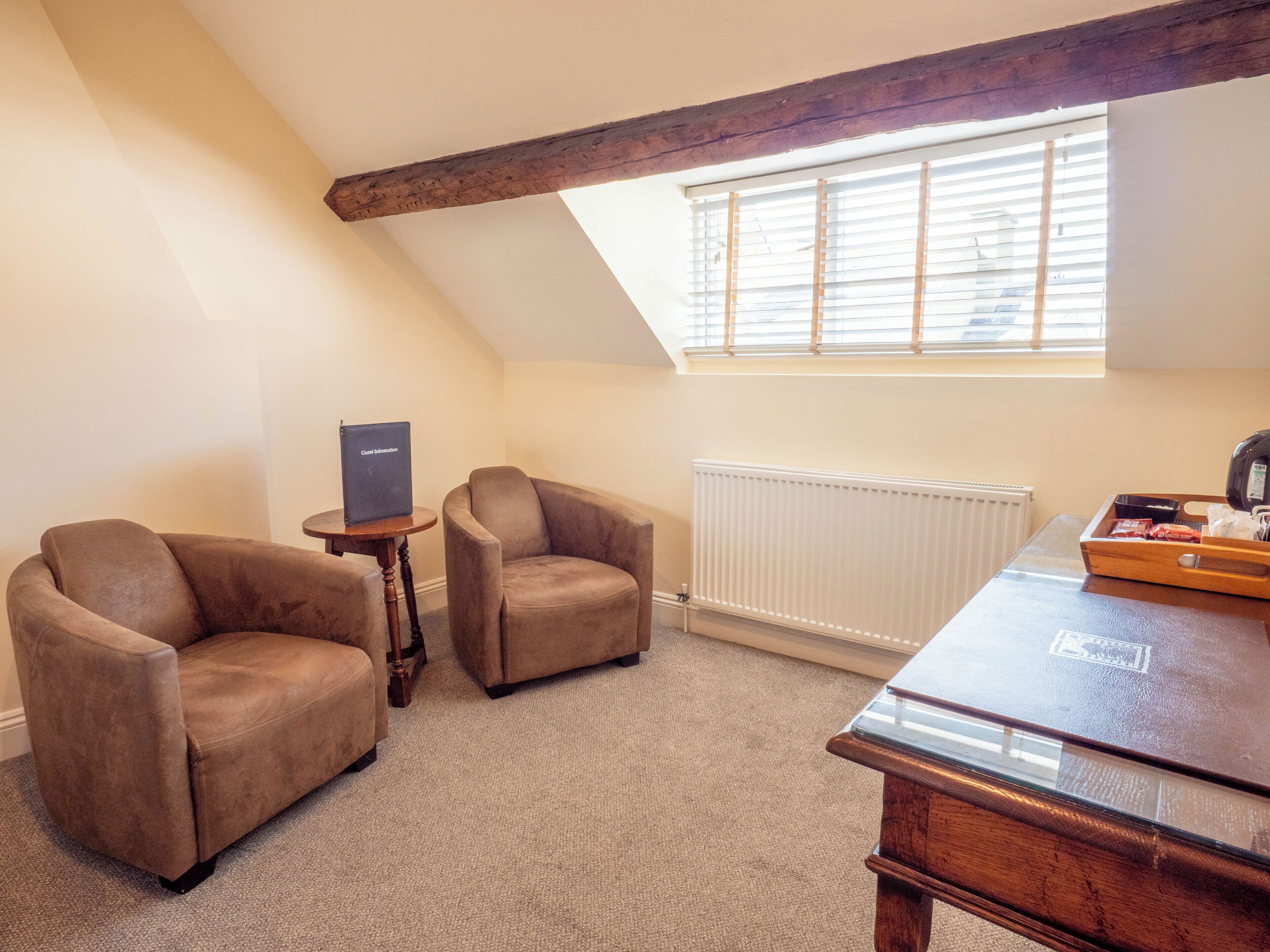 A room with two brown leather armchairs, a small wooden table with a laptop, a wooden desk with a tray of items, a radiator, and a window with blinds.