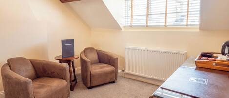A room with two brown leather armchairs, a small wooden table with a laptop, a wooden desk with a tray of items, a radiator, and a window with blinds.