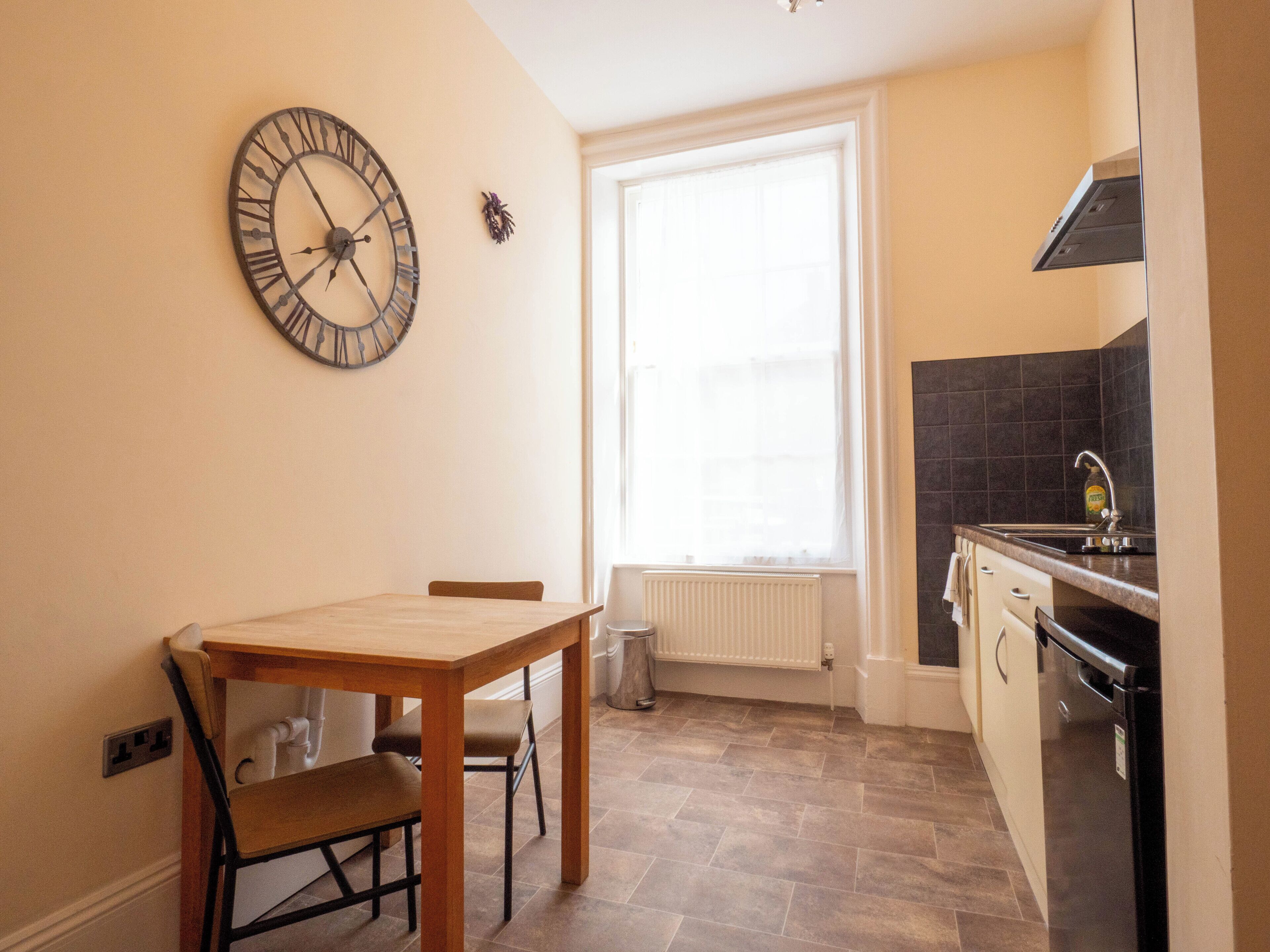 A small kitchen with a wooden table, a clock, a window, and a sink.