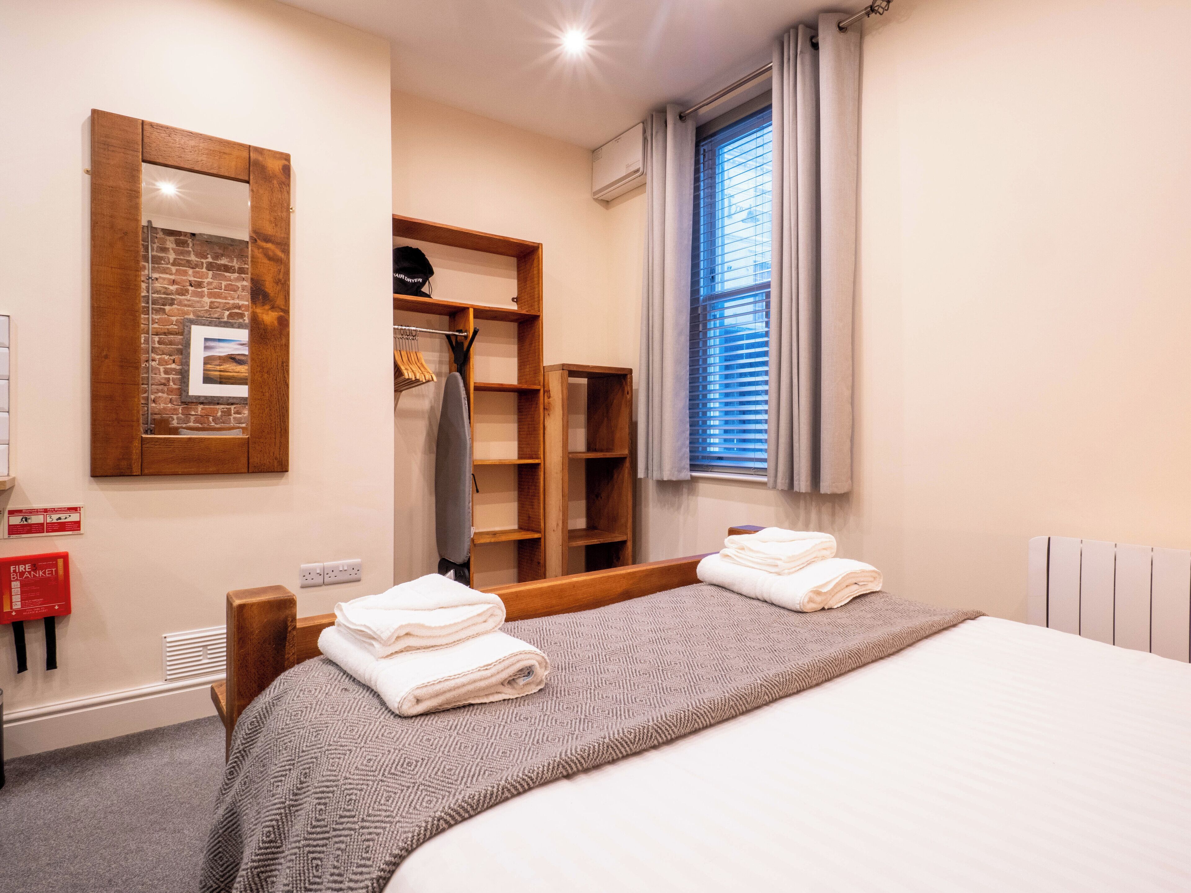 Habitación de hotel con cama, armario, espejo, ventana con persianas y señal de manta ignífuga.