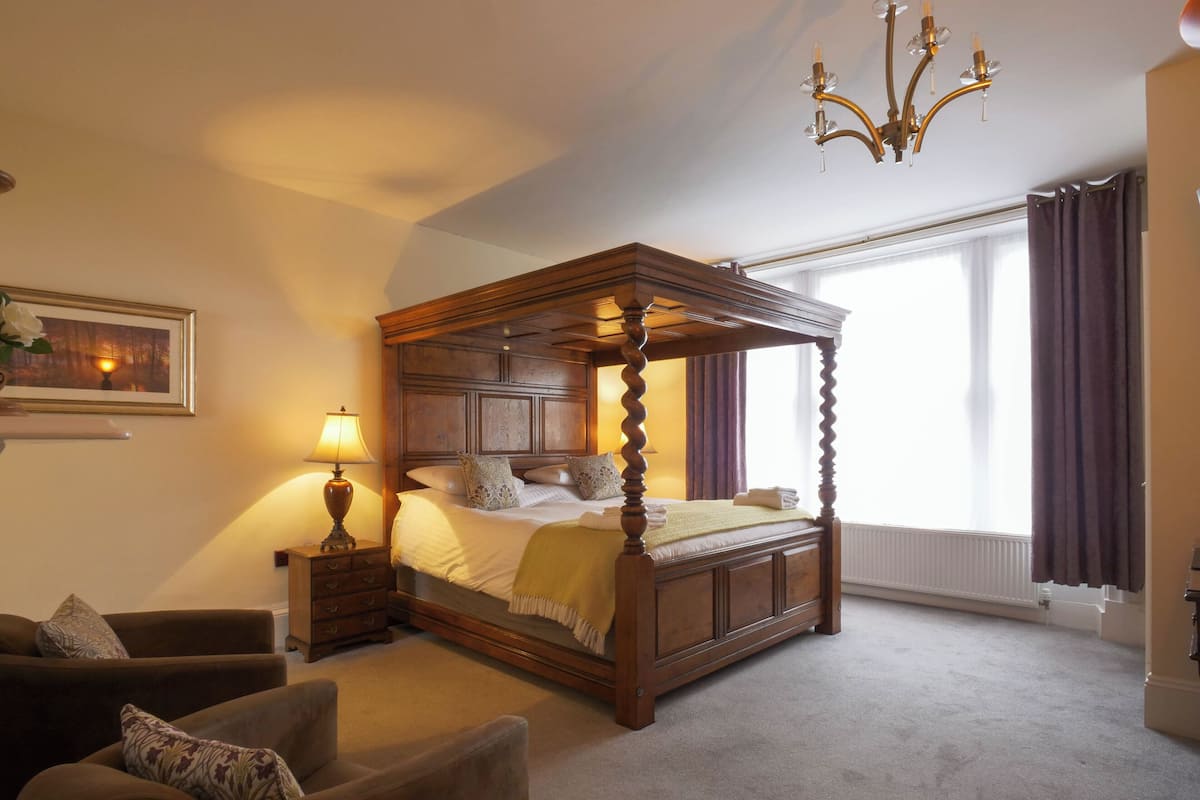 Ein Bett mit Baldachin und einem hölzernen Kopfteil, ein Nachttisch mit Lampe, ein brauner Sessel und ein großes Fenster mit Vorhängen.