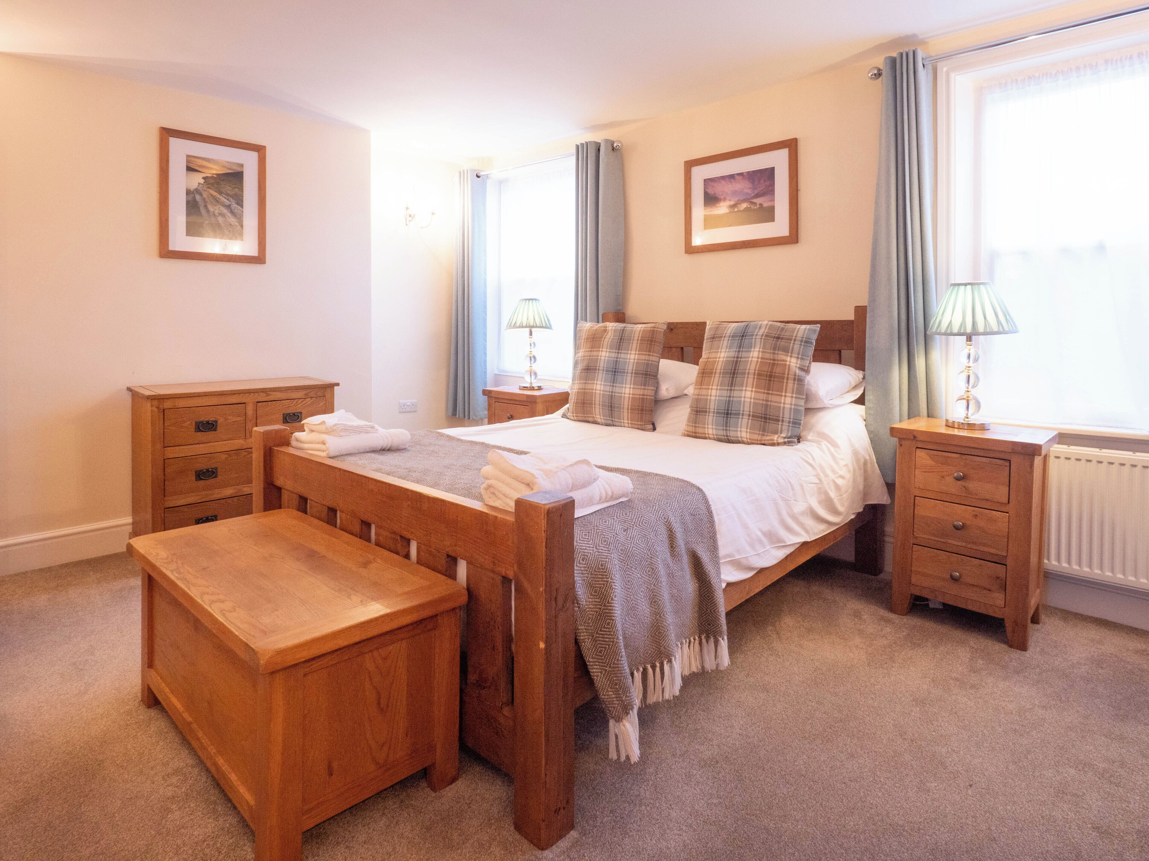 Un dormitorio con cama y banco de madera, mesita de noche, lámpara y ventana con cortinas.