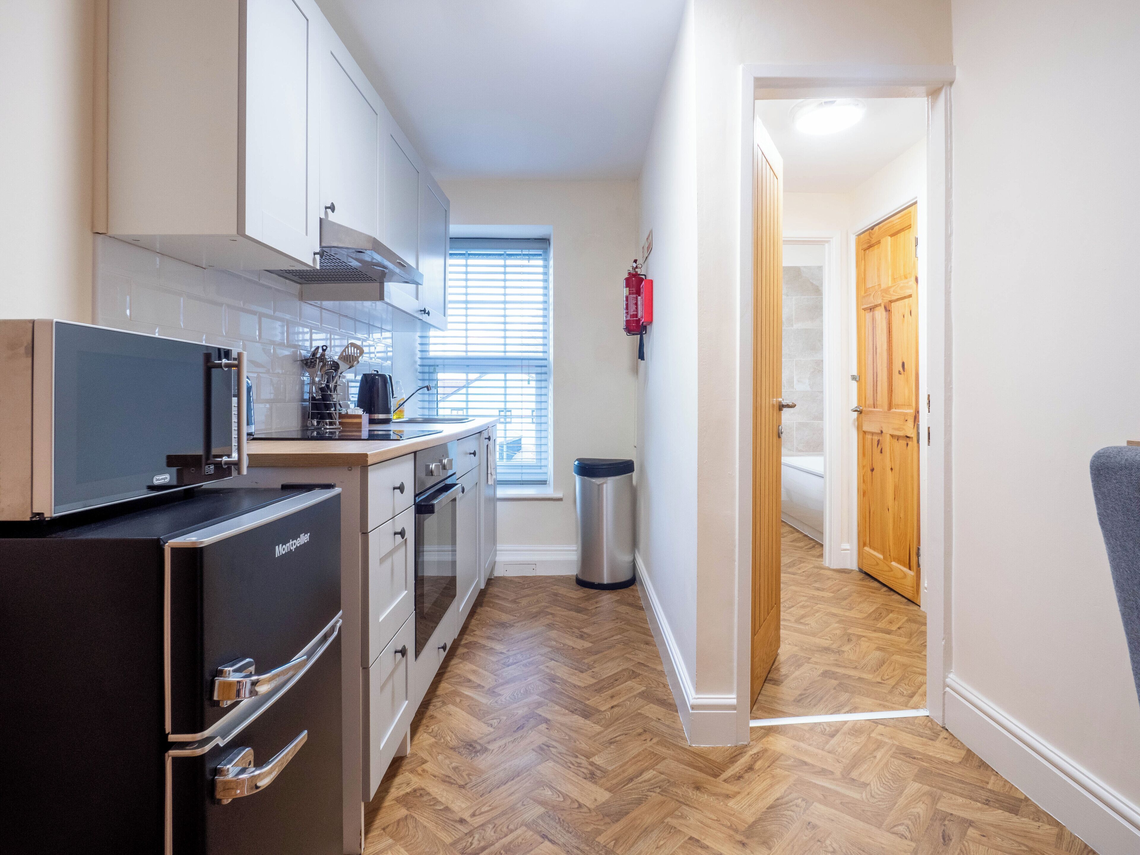 Una cocina compacta con electrodomésticos modernos, una ventana con persianas y una puerta de madera que da a otra habitación.