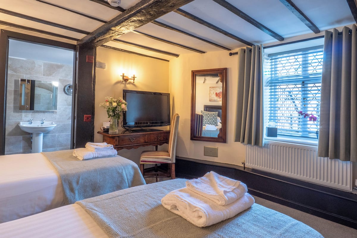 Ein Hotelzimmer mit zwei Betten, einem Fernseher, einem Schreibtisch mit Blumenvase, einem Spiegel und einem Fenster mit Vorhängen.