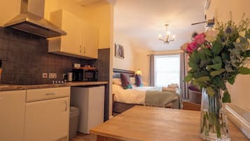 A compact hotel room with a kitchenette, a bed with a headboard, a bedside table, and a vase with flowers.
