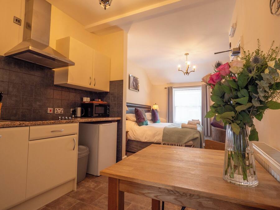 A compact hotel room with a kitchenette, a bed with a headboard, a bedside table, and a vase with flowers.