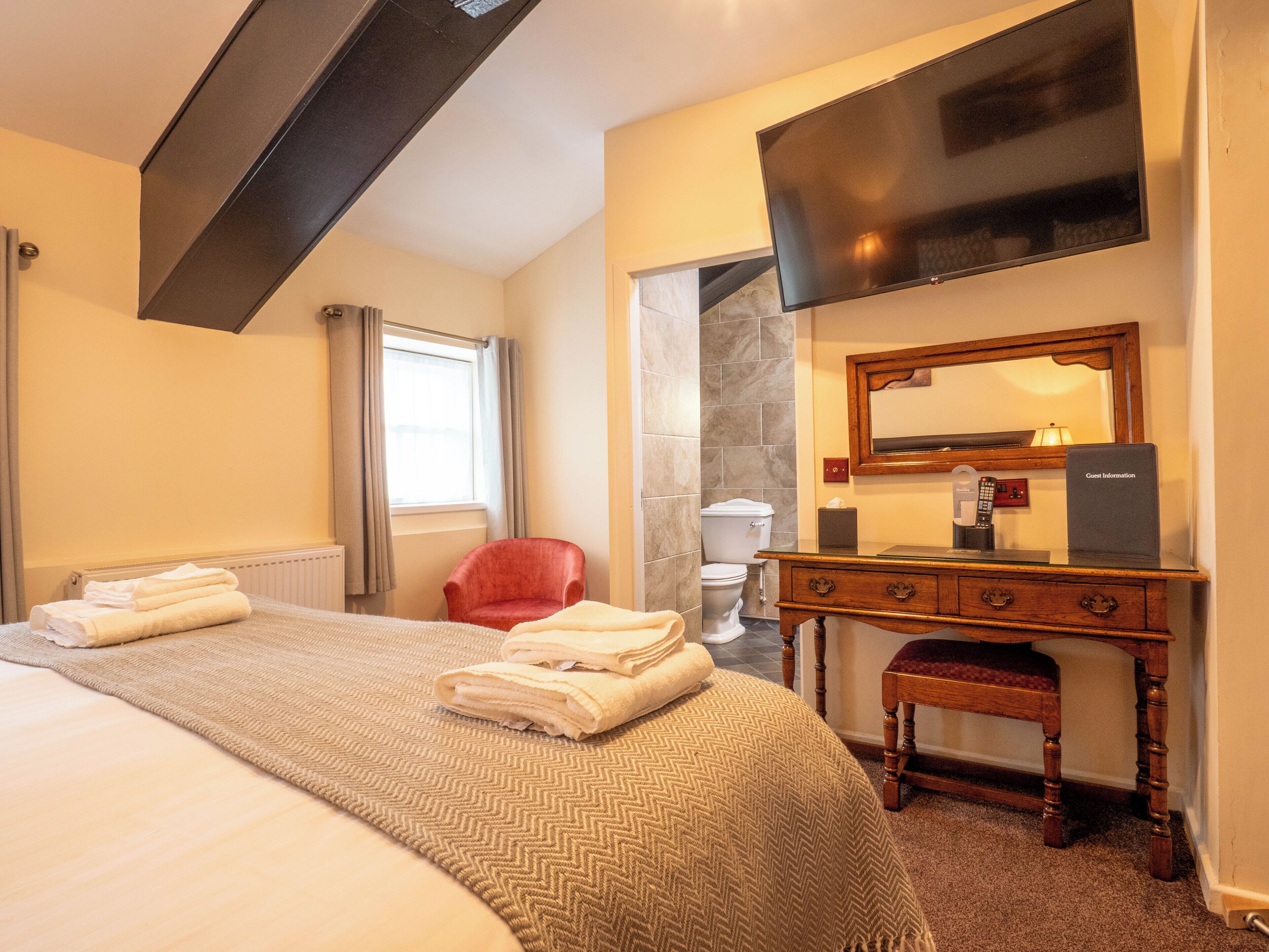 Een hotelkamer met een bed, een rode stoel, een houten dressoir, een flatscreen televisie en een spiegel.