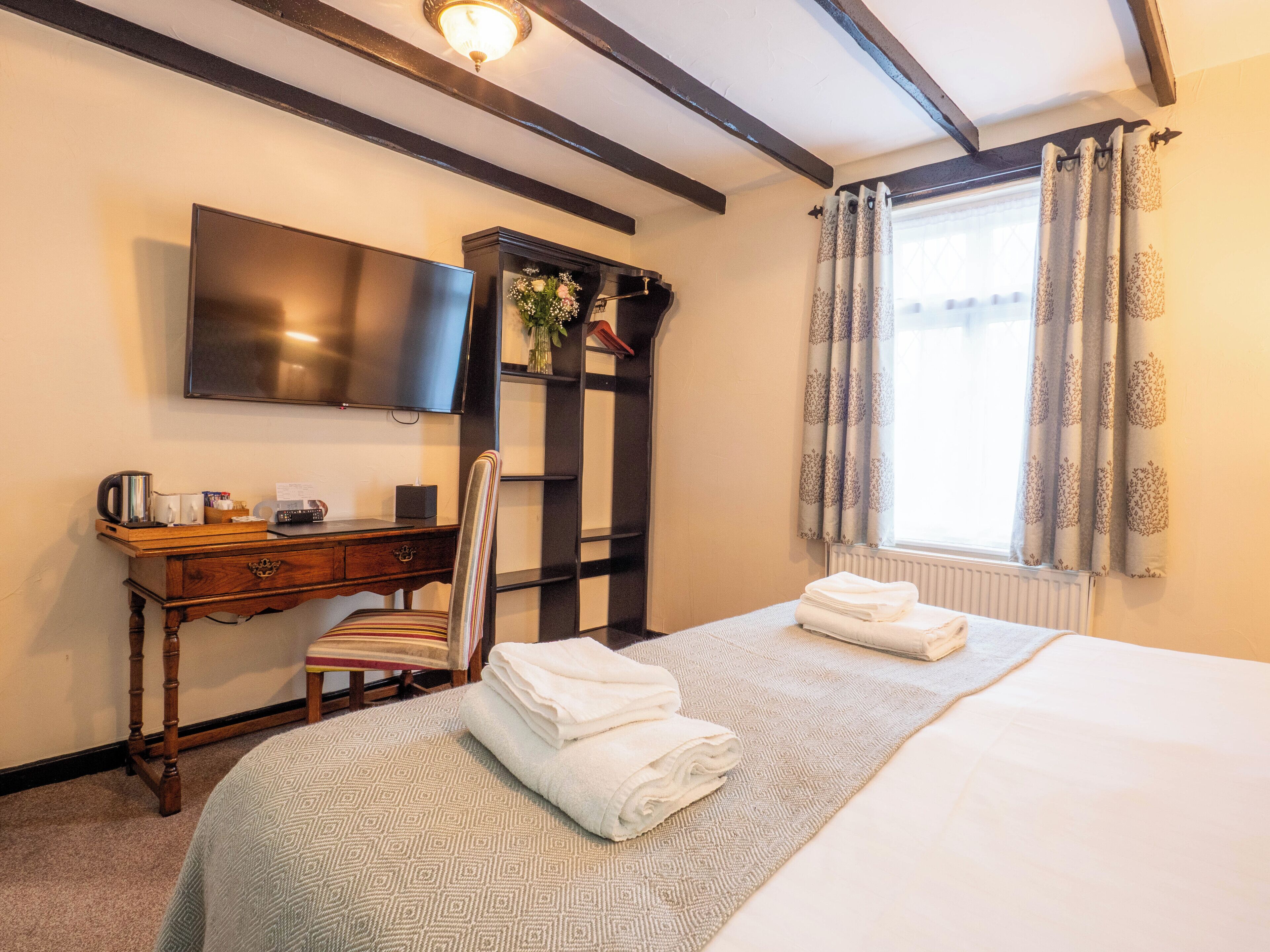 Ein Hotelzimmer mit Bett, Schreibtisch, Flachbildfernseher, Stuhl, Ablagefläche mit Vase und einem Fenster mit Vorhängen.