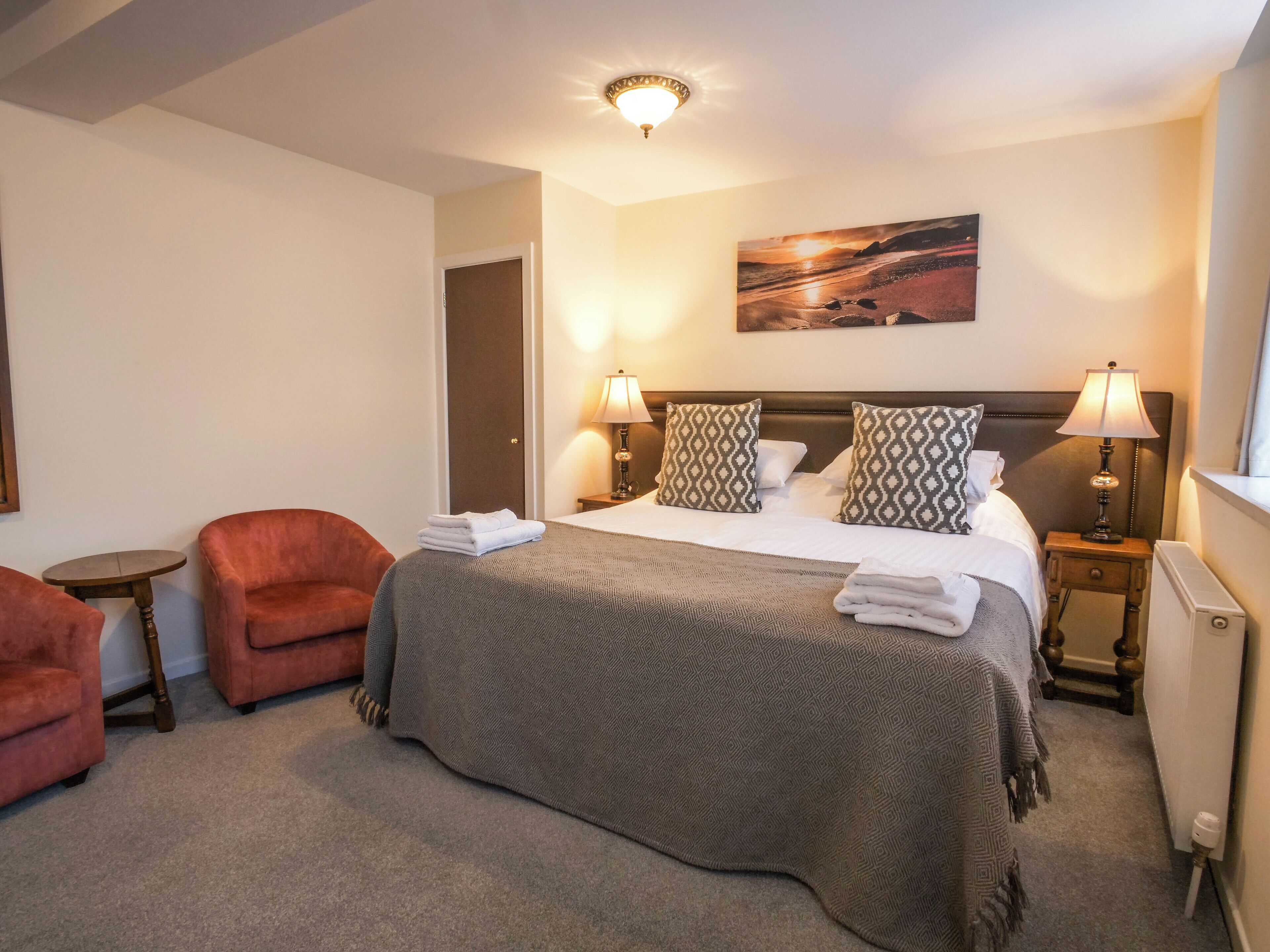 A hotel room with a bed, two bedside lamps, a painting on the wall, a door, and two armchairs.