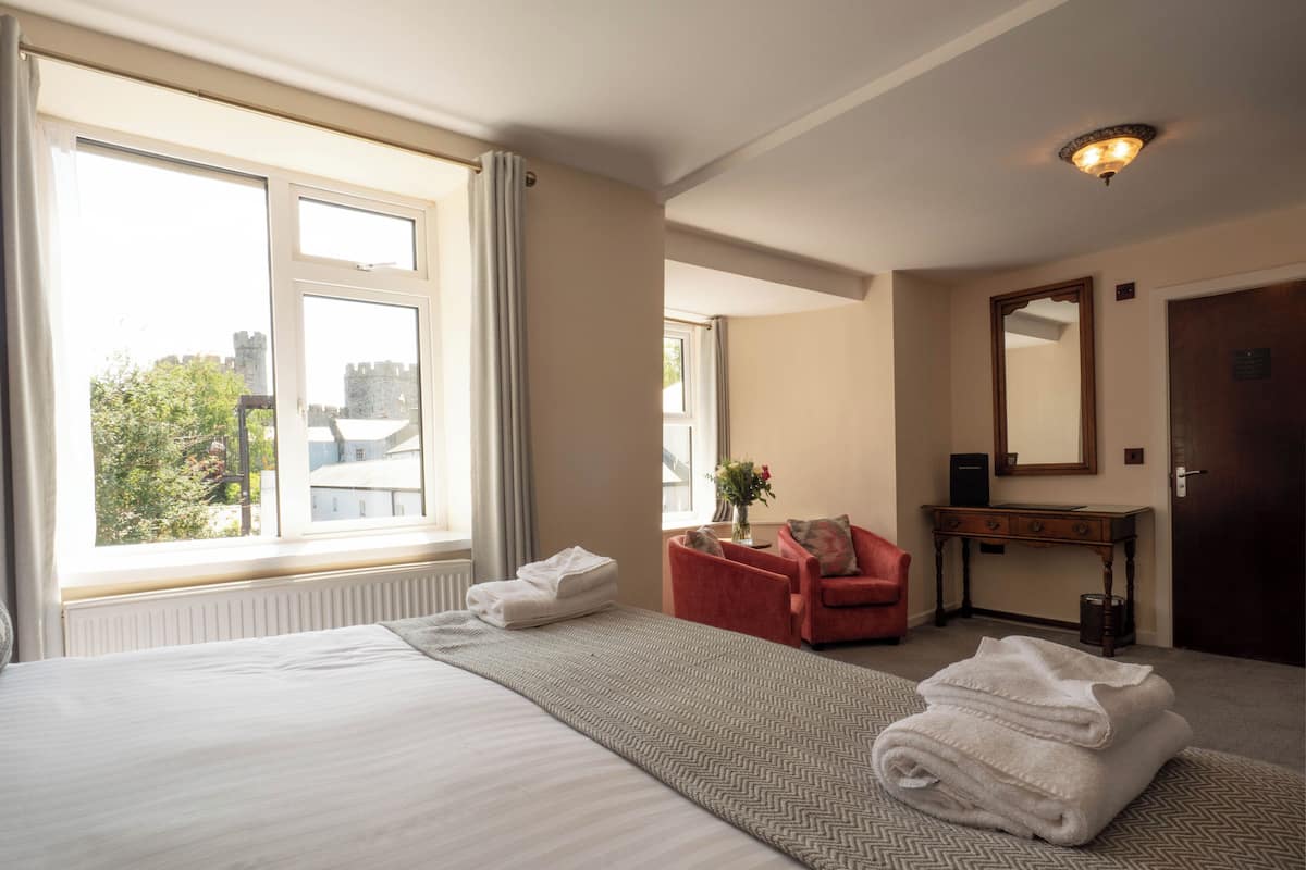 Ein Hotelzimmer mit einem großen Bett, einer Sitzecke mit einem Sessel, einem kleinen Tisch mit einer Vase und einem Spiegel an der Wand.